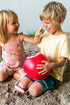 Two children playing on a sandy beach with a red ball shaped bucket