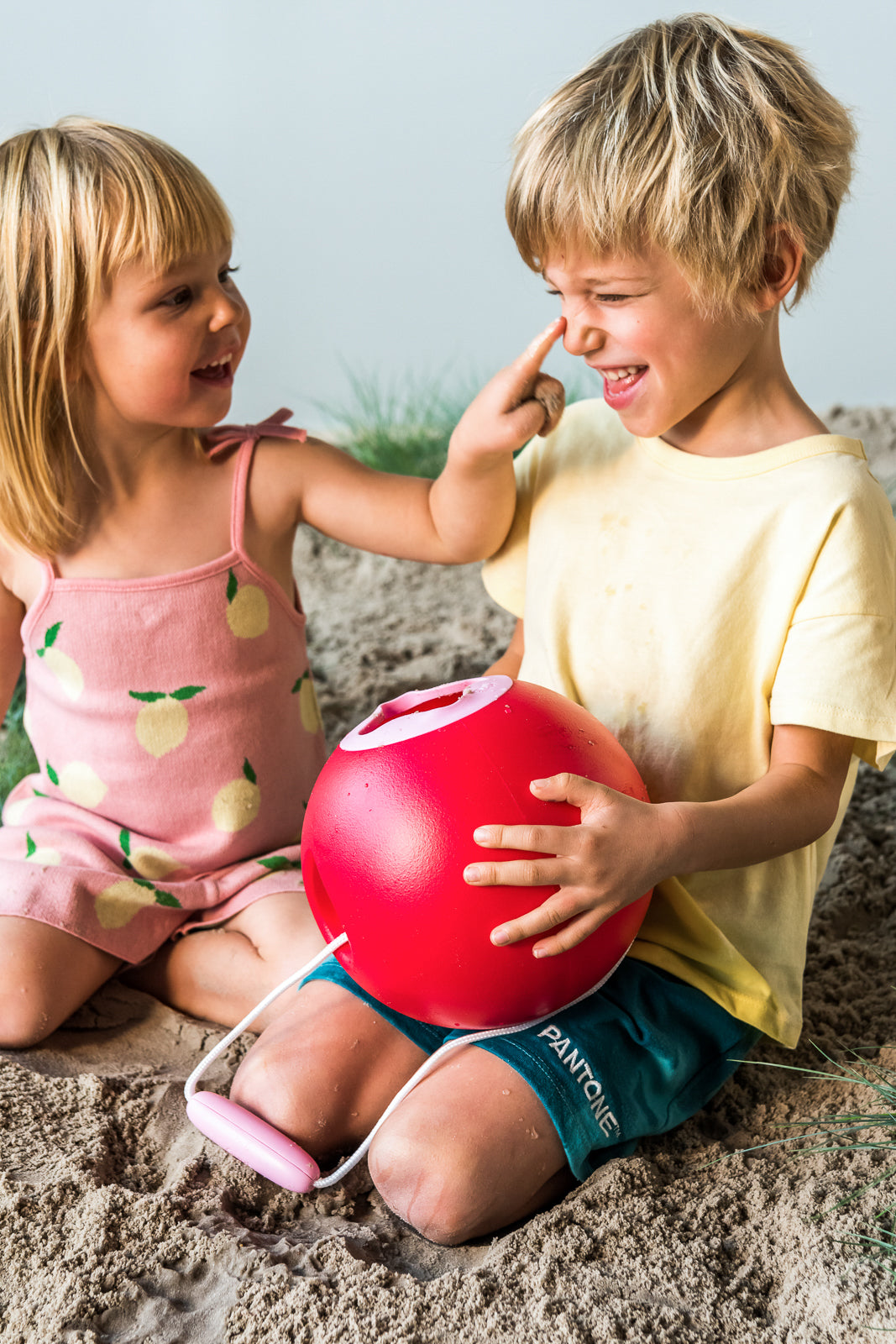 Two children playing on a sandy beach with a red ball shaped bucket