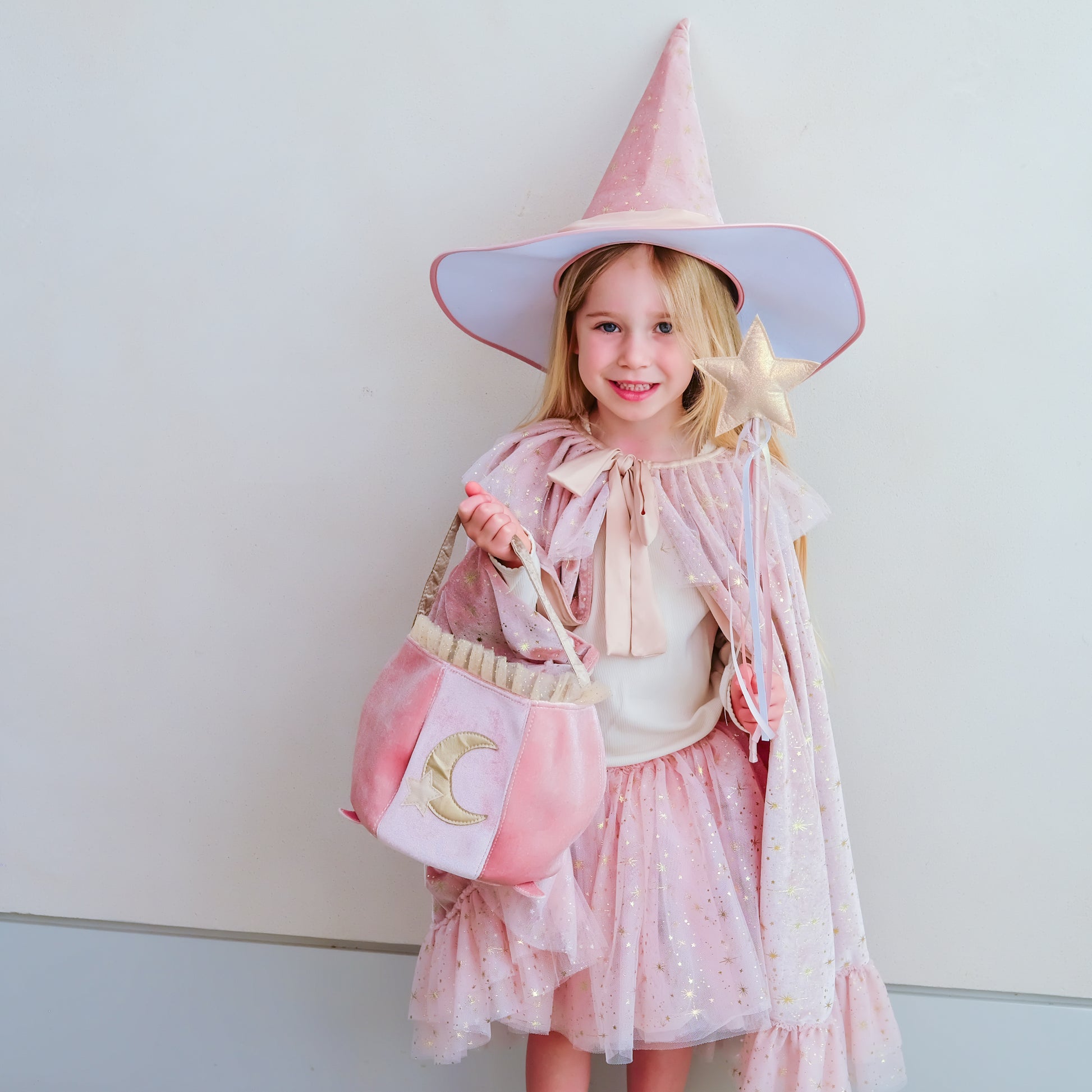 A young girl wearing a pink witch costume set, including a pointed hat, cape, dress, and matching bag with a crescent moon design, standing against a plain light gray wall.