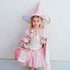 A young girl wearing a pink wizard costume stands against a plain light gray wall, holding a pink star wand and a matching pink moon bag.