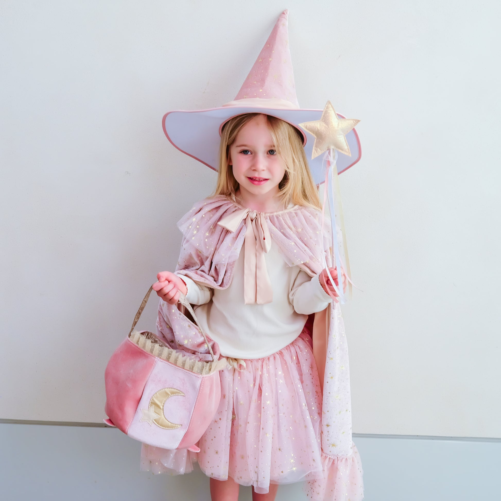 A young girl wearing a pink wizard costume stands against a plain light gray wall, holding a pink star wand and a matching pink moon bag.