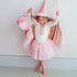 A young girl wearing a pink and white witch costume, including a pointed hat, cape, and tutu skirt, posing against a plain light gray wall.