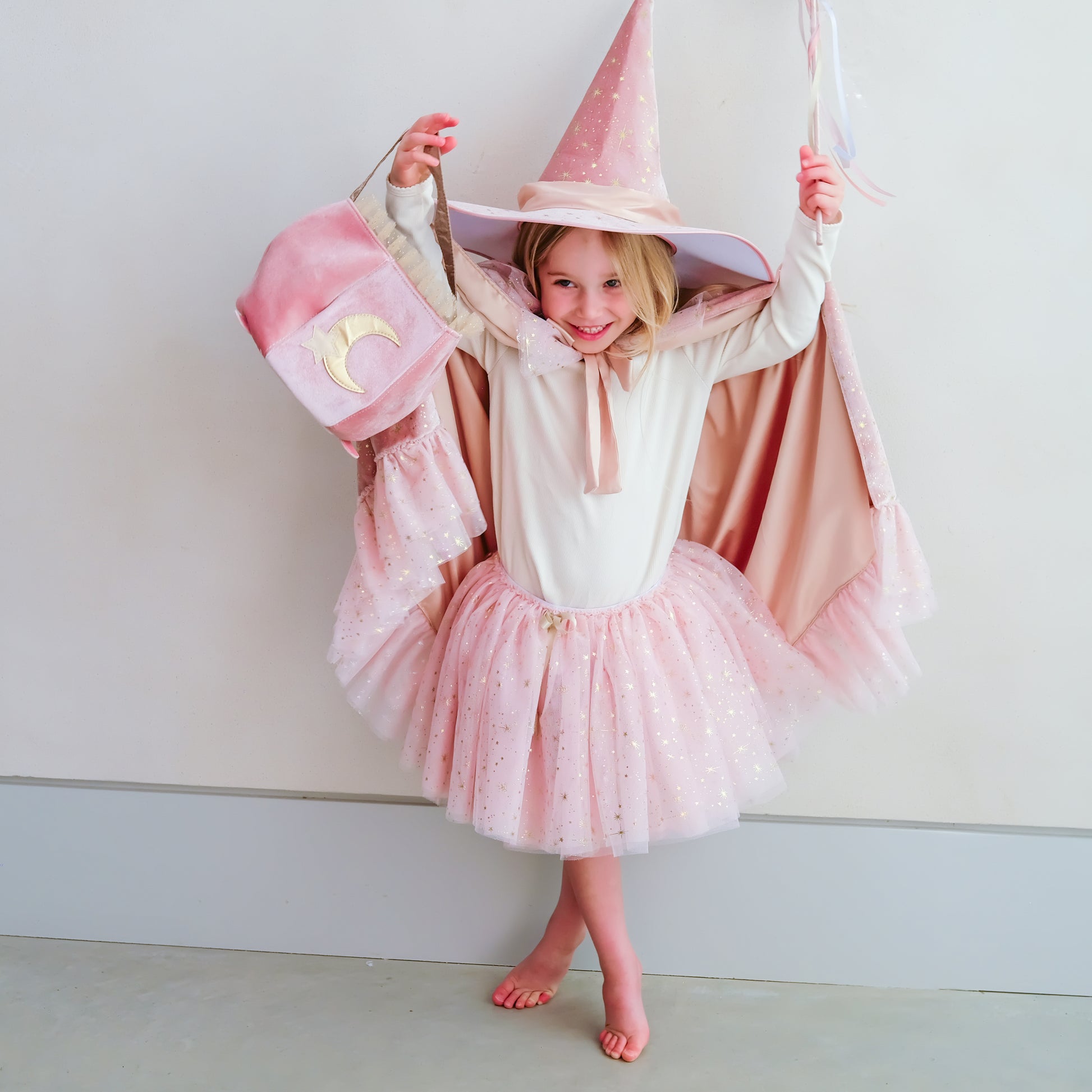A young girl wearing a pink and white witch costume, including a pointed hat, cape, and tutu skirt, posing against a plain light gray wall.