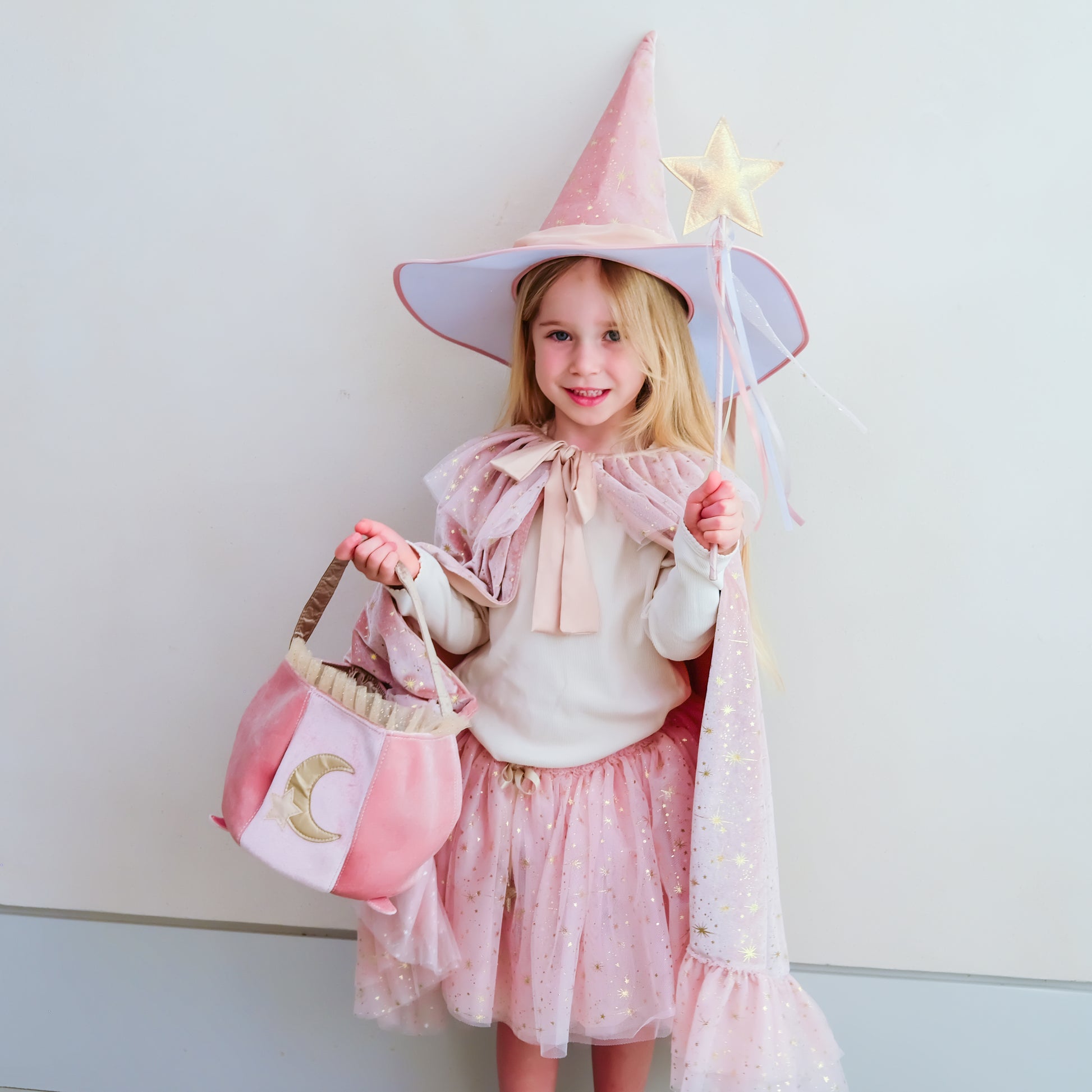 A young girl wearing a pink witch costume with a pointed hat, cape, and skirt, holding a pink purse with a gold crescent moon and a wand with a star, standing against a plain light gray wall.