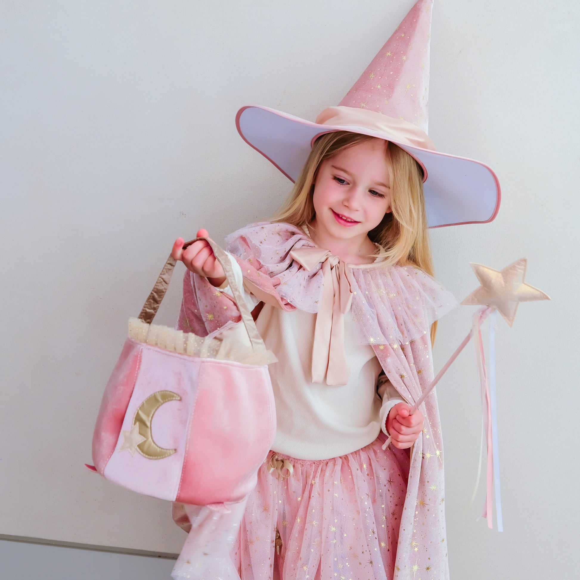 A young girl wearing a pink witch costume with a pointed hat, star wand, and a pink bucket bag with a gold crescent moon, standing against a plain light gray wall.