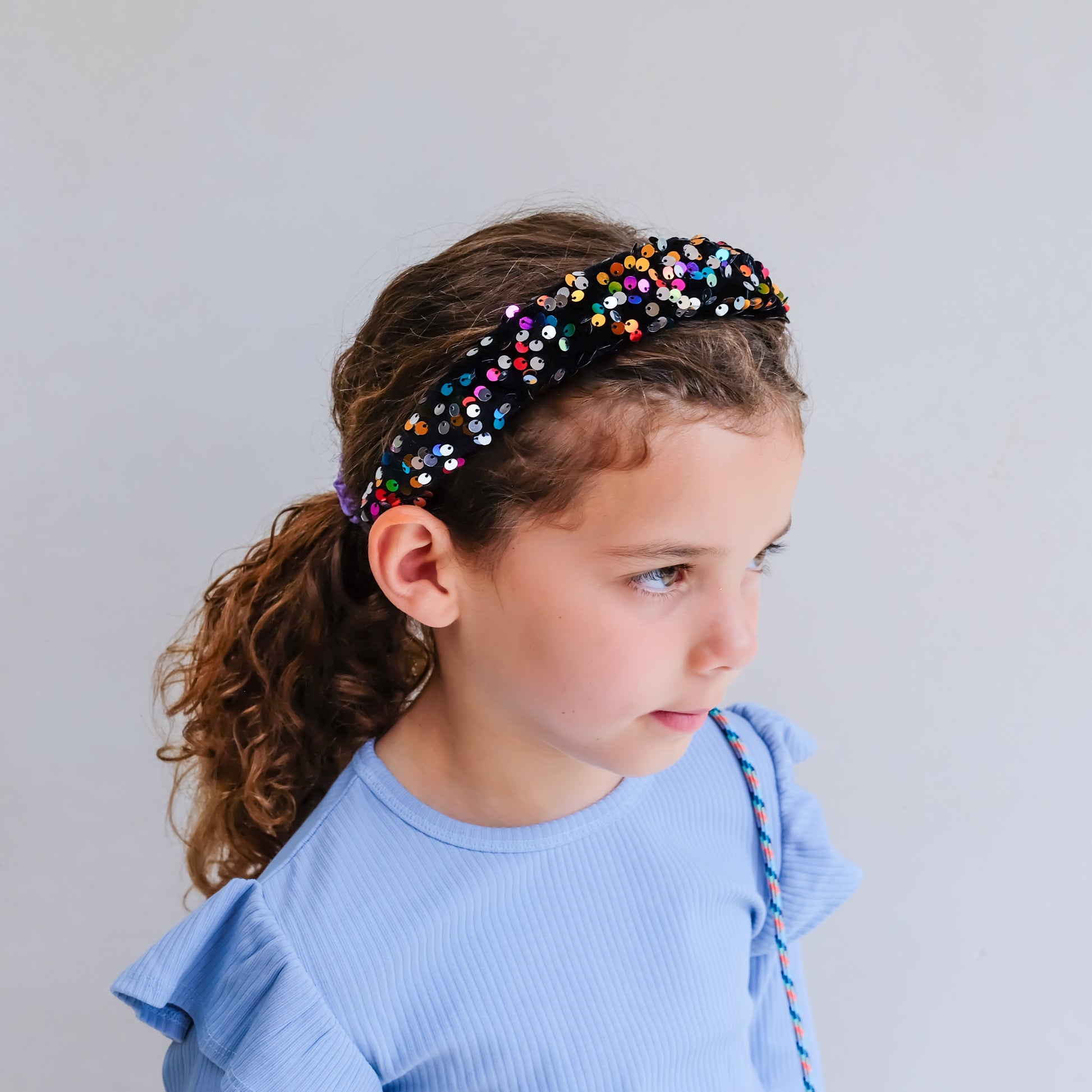 A young girl with curly brown hair wearing a black headband adorned with colorful rhinestones, paired with a light blue ruffled top, shown in profile against a plain light gray background.