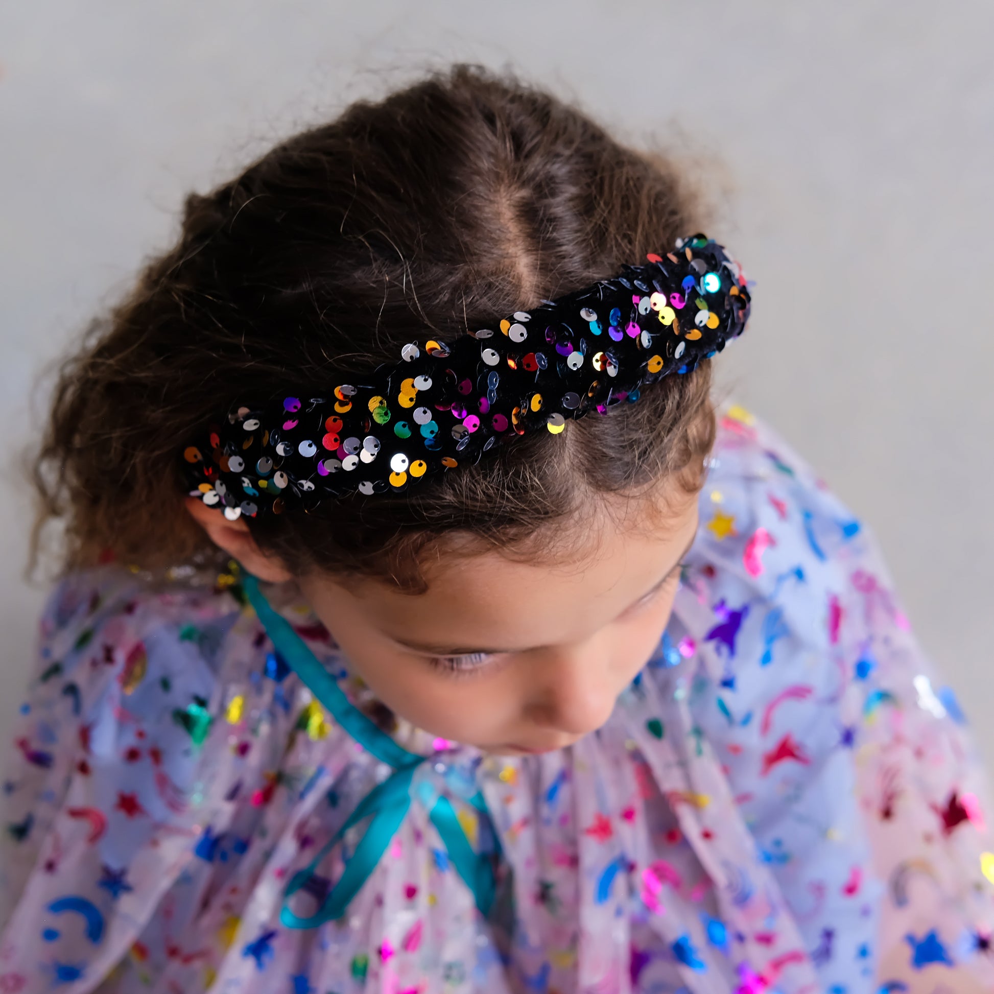A close-up lifestyle shot of a young girl with curly brown hair wearing a black sequined headband and a light purple tulle dress covered in colorful sequins.