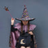 A child dressed as a witch wearing a tall black hat covered in multicolored sequins, a pink ruffled collar, a black dress, and a matching sequined cape, holding a star wand and a black bucket with a gold crescent moon and star.