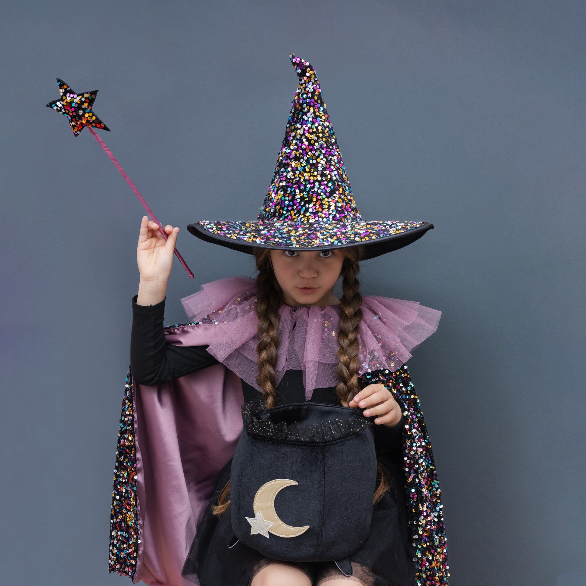 A child dressed as a witch wearing a tall black hat covered in multicolored sequins, a pink ruffled collar, a black dress, and a matching sequined cape, holding a star wand and a black bucket with a gold crescent moon and star.