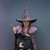 A child wearing a sparkly multicolor witch costume hat, a pink tulle collar, a black dress, and a glittery cape, holding a pink wand and a black bucket with a gold crescent moon, against a dark gray background.