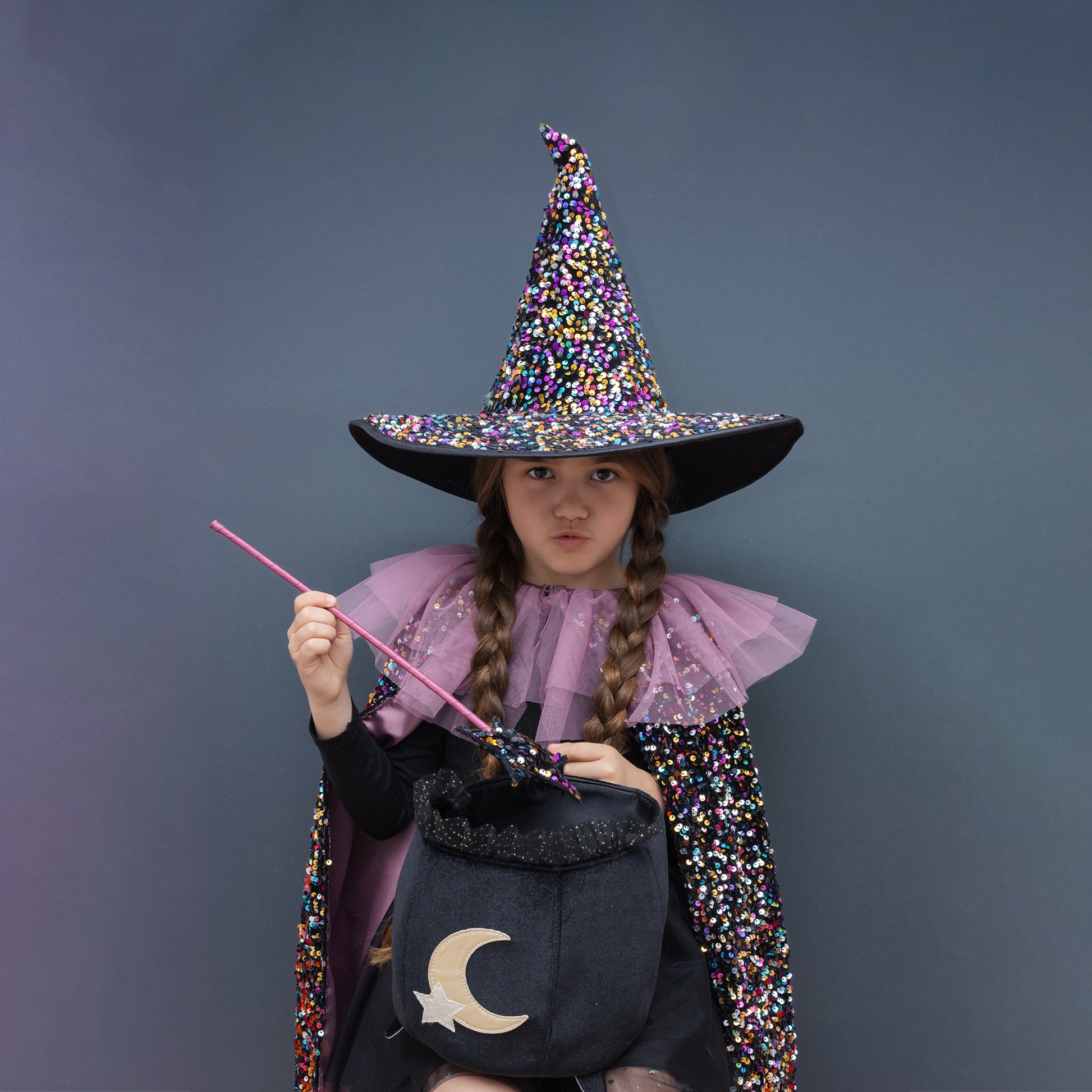 A child wearing a sparkly multicolor witch costume hat, a pink tulle collar, a black dress, and a glittery cape, holding a pink wand and a black bucket with a gold crescent moon, against a dark gray background.