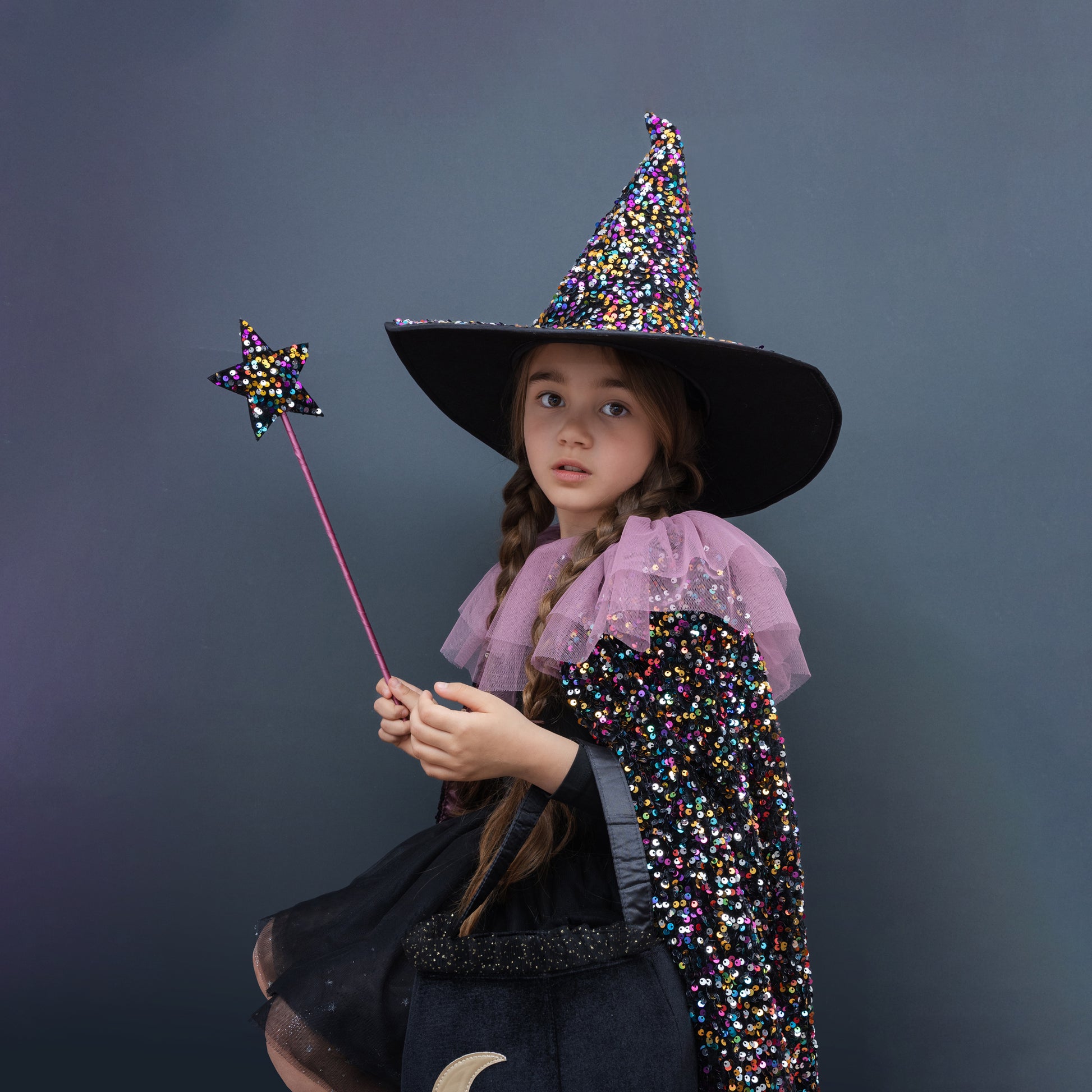 Child in a Halloween witch costume with a colorful hat and wand against a dark background