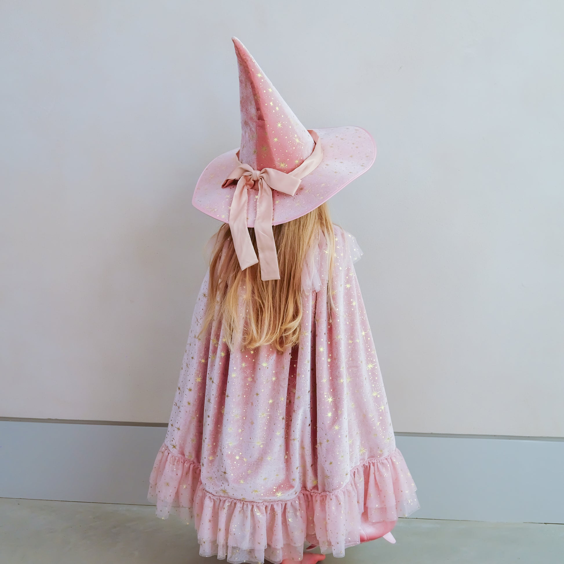 A young girl viewed from behind wearing a pink witch hat with a matching pink cape. The fabric is light pink with tiny white polka dots and subtle glitter, and the hat is adorned with a large pink bow.