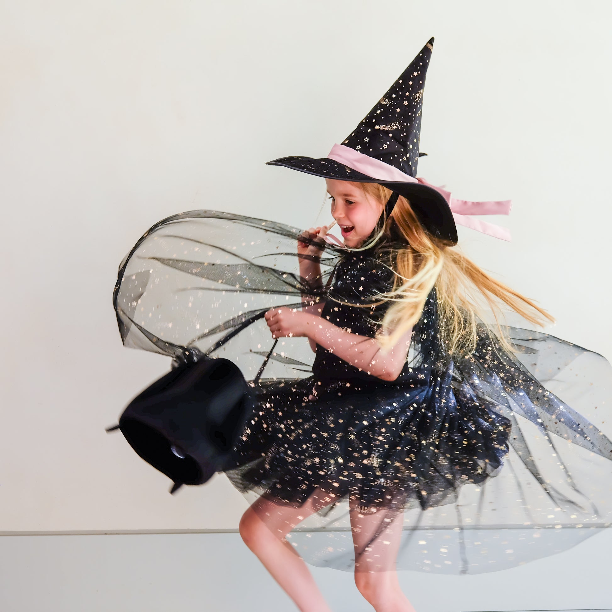 A young girl wearing a black witch costume with a black hat, pink ribbon, and sheer black tulle skirt, running while holding a black bucket against a plain light background.
