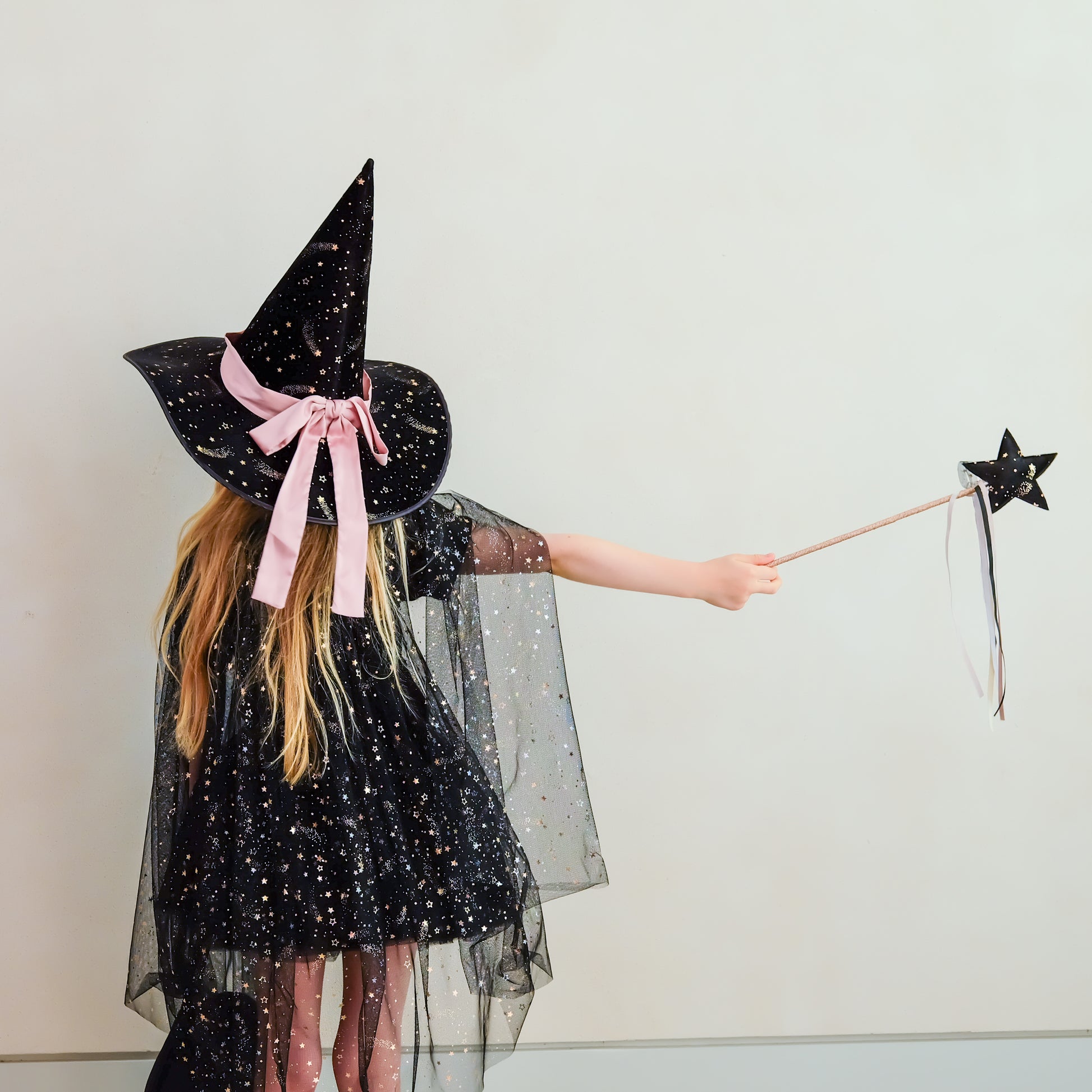 A child wearing a black glittery witch costume with a large pointed hat, pink ribbon bow, sheer black cape, and holding a black star wand against a plain light background.