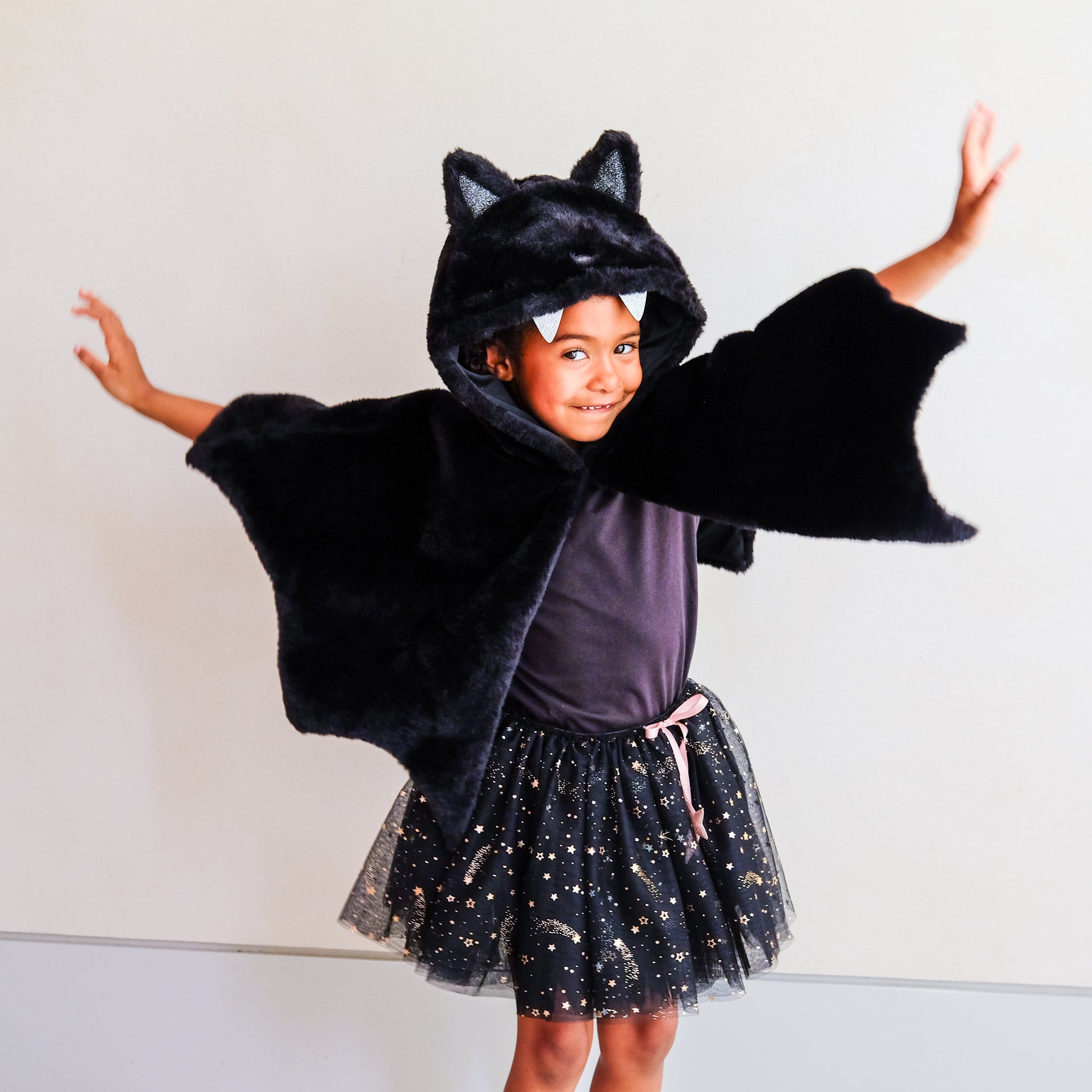 A young child wearing a black plush bat costume cape with attached hood and ears, posed with arms outstretched against a plain light background.