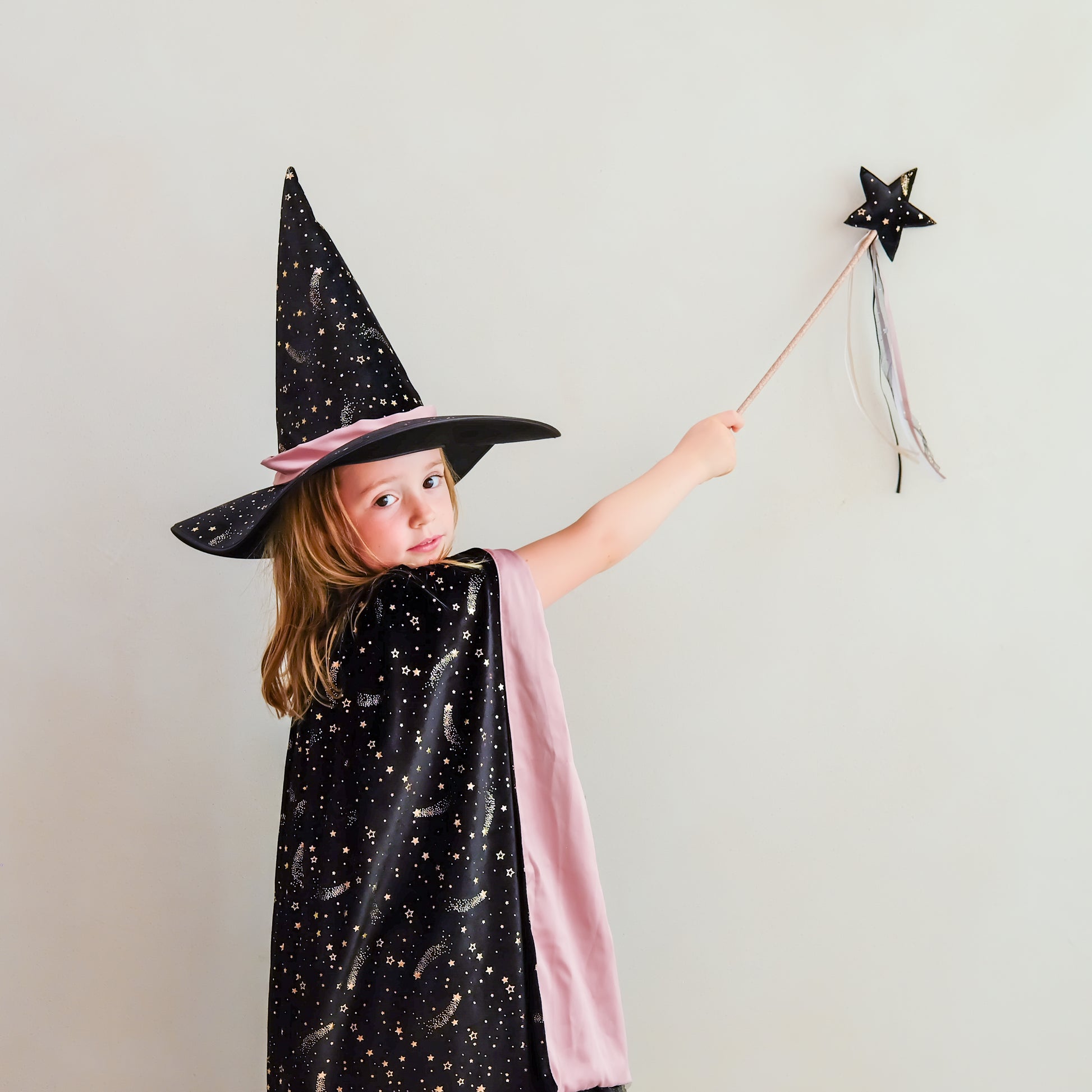 A young girl wearing a black witch hat with gold stars and a matching black cape with gold moon patterns, draped in a pink fabric, holding a black star wand against a plain light gray background.