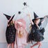 Three children wearing witch costumes and hats pose together against a plain light gray wall. One child wears a black dress with gold star confetti and a pink hat band, another wears a pink dress with a pink witch hat, and the third wears a black dress with gold star confetti and a black hat.