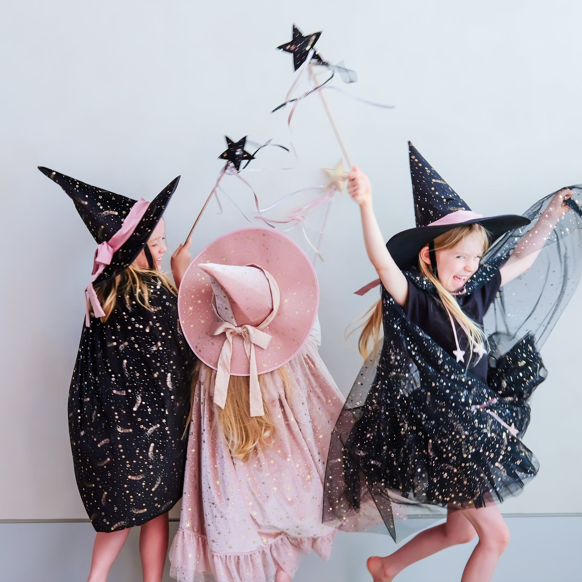 Three children wearing witch costumes and hats pose together against a plain light gray wall. One child wears a black dress with gold star confetti and a pink hat band, another wears a pink dress with a pink witch hat, and the third wears a black dress with gold star confetti and a black hat.
