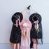 Three children wearing witch-themed costumes stand against a plain light gray wall, holding a small wand with stars. The child on the left wears a black dress with gold speckles and a black witch hat with a pink ribbon.