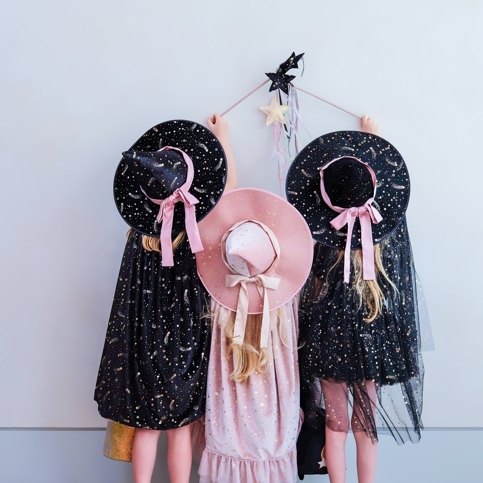 Three children wearing witch-themed costumes stand against a plain light gray wall, holding a small wand with stars. The child on the left wears a black dress with gold speckles and a black witch hat with a pink ribbon.