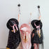 Three children wearing witch costumes stand against a plain light wall, each holding a black star wand. The child on the left wears a black dress with gold speckles and a large black hat with a pink ribbon.