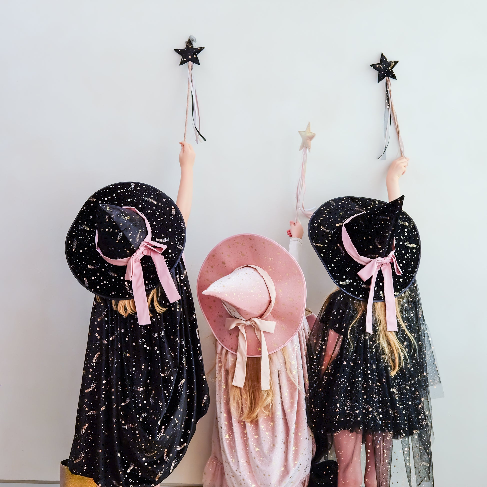 Three children wearing witch costumes stand against a plain light wall, each holding a black star wand. The child on the left wears a black dress with gold speckles and a large black hat with a pink ribbon.