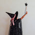 A child wearing a black glittery witch costume with a pointed hat and pink ribbon bow, holding a black star wand against a plain light background.