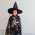 A young child wearing a black witch costume with gold star details, a tall pointed witch hat, and a sheer black cape with pink ribbons, holding a silver and black toy against a plain light gray wall.