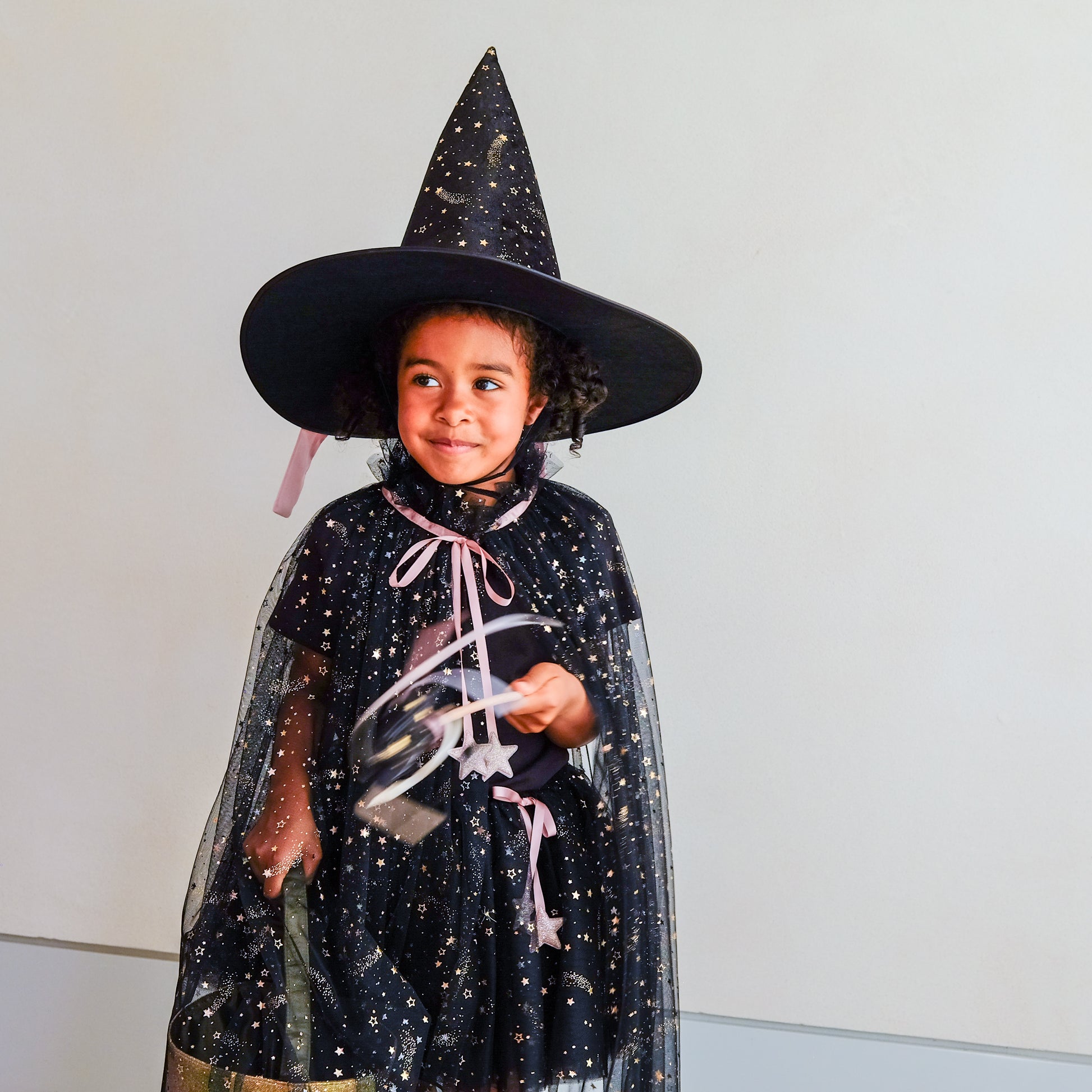 A young child wearing a black witch costume with gold star details, a tall pointed witch hat, and a sheer black cape with pink ribbons, holding a silver and black toy against a plain light gray wall.