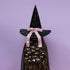 A person wearing a tall black witch hat with gold speckles, a pink satin bow, and a matching black cape covered in colorful hair clips, set against a solid lavender background.