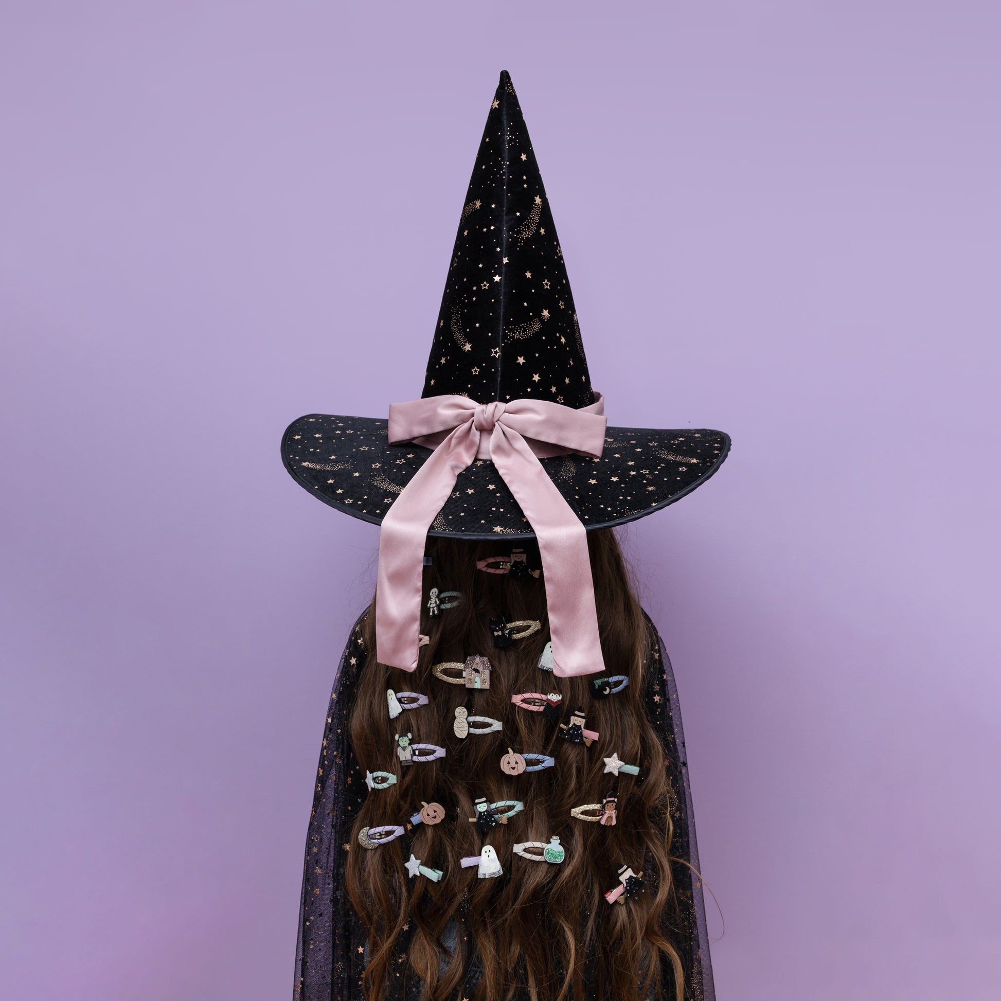 A person wearing a tall black witch hat with gold speckles, a pink satin bow, and a matching black cape covered in colorful hair clips, set against a solid lavender background.
