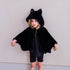 A young girl wearing a black plush bat cape costume with a hood featuring cat ears, posed against a plain light gray wall.