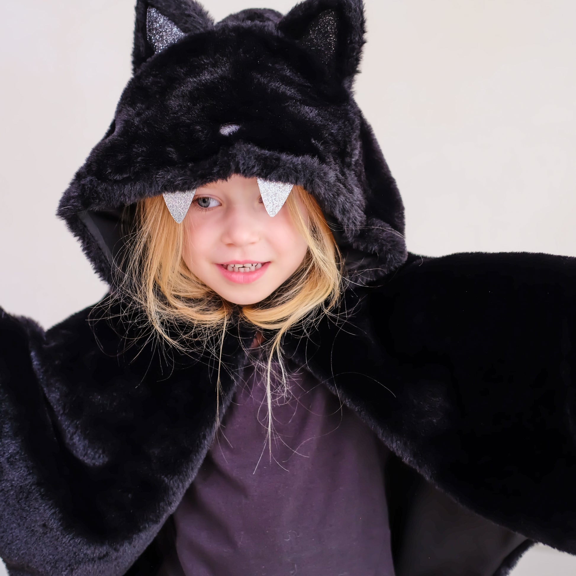 A young girl wearing a black plush bat costume cape with attached hood, cat ears, and silver glitter bat wings peeking out from under the hood, photographed against a plain light background.