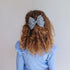 A child with long, curly light brown hair seen from behind, wearing a large gray and white patterned bow hair clip. The child is wearing a light blue ribbed top against a plain light gray background.