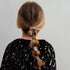 Back view of a child with long light brown hair styled in a low ponytail, wearing three small decorative hair clips, including a fairy clip and a moon clip, against a plain light gray background.
