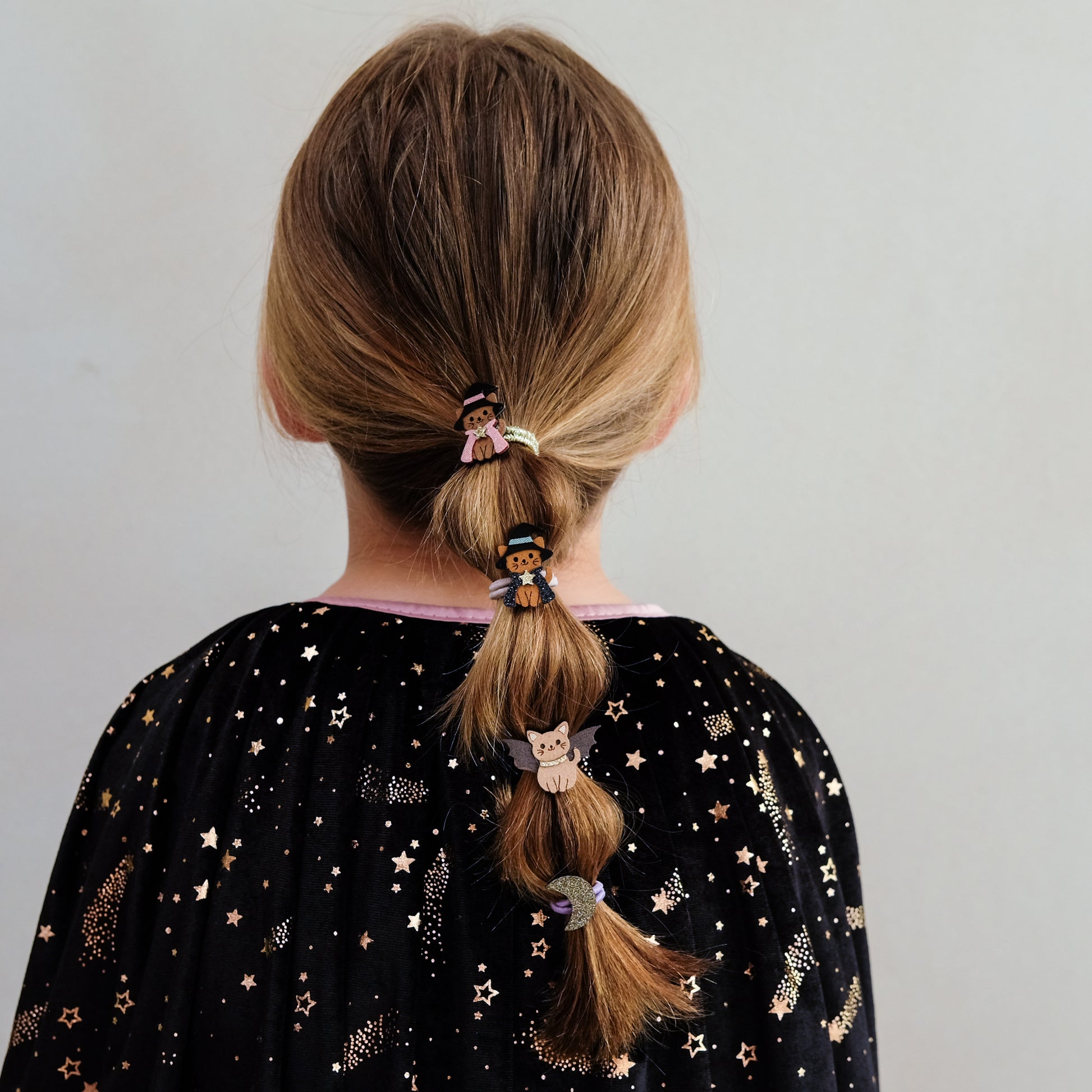 Back view of a child with long light brown hair styled in a low ponytail, wearing three small decorative hair clips, including a fairy clip and a moon clip, against a plain light gray background.