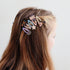 A close-up side view of a person with long brown hair wearing a set of colorful decorative hair clips attached to a hair tie. The clips feature whimsical cartoon and animal designs in pastel and bright colors against a plain light gray background.