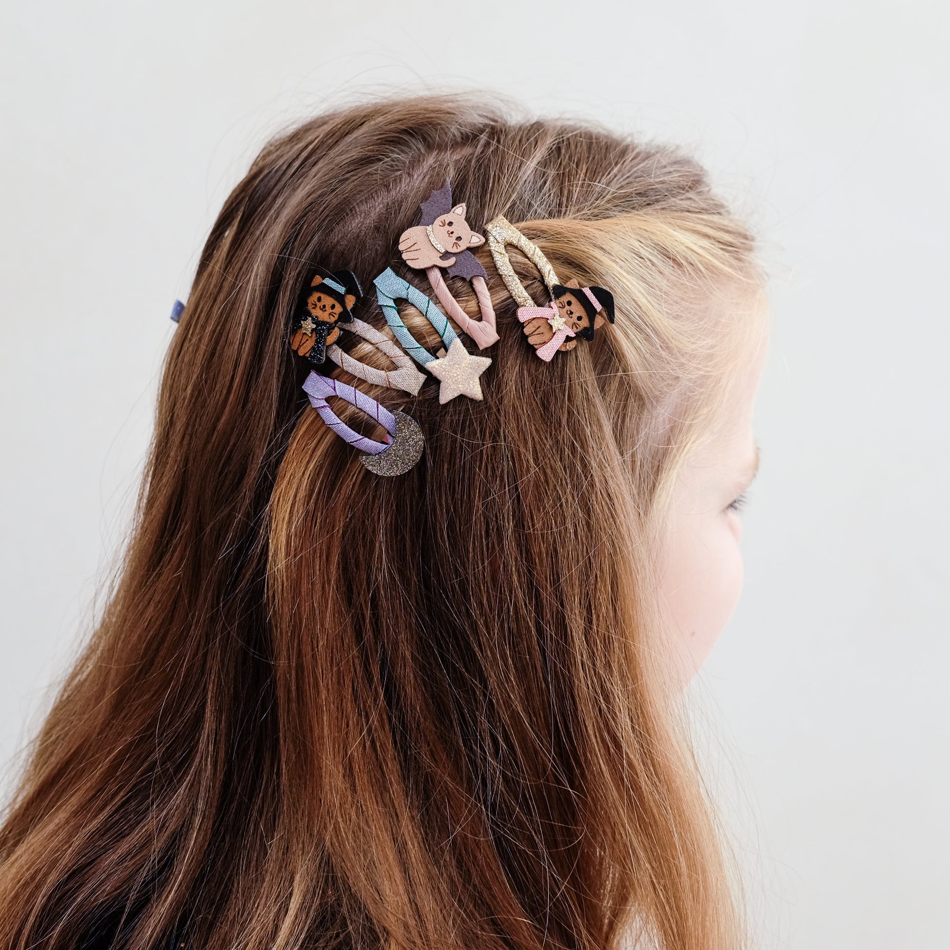 A close-up side view of a person with long brown hair wearing a set of colorful decorative hair clips attached to a hair tie. The clips feature whimsical cartoon and animal designs in pastel and bright colors against a plain light gray background.