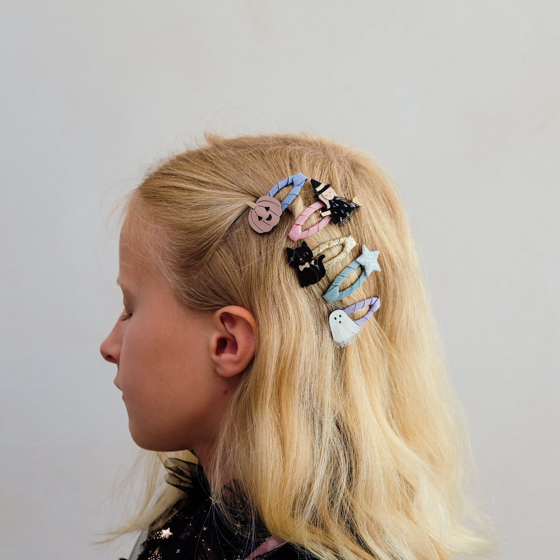 A side-profile view of a young girl with blonde hair wearing a set of colorful, whimsical hair clips, including a button, a ghost, a star, and a small cat, against a plain light gray background.