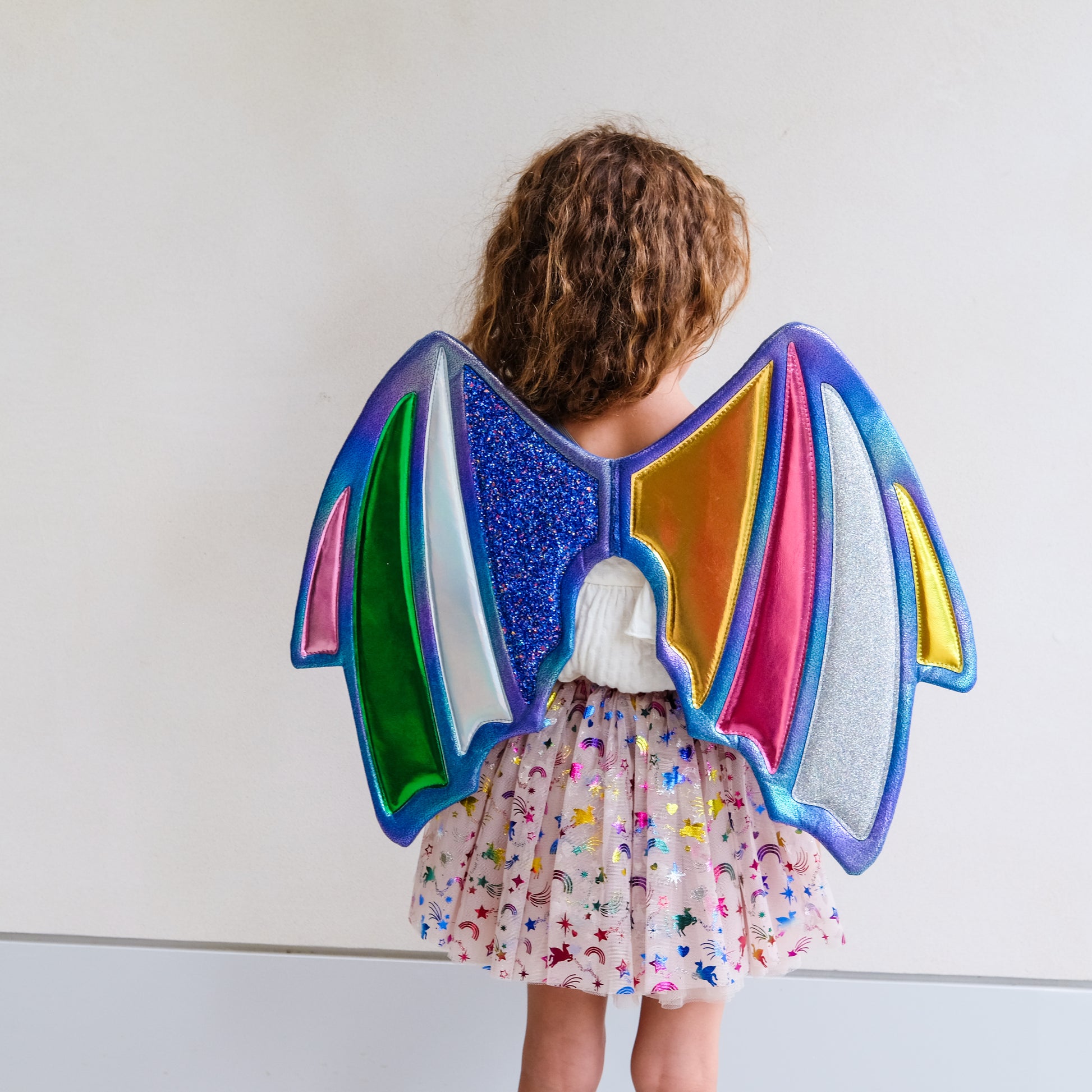 A child viewed from behind wearing a colorful fairy wing costume accessory with glittery blue, pink, green, yellow, orange, and silver panels, paired with a light pink floral skirt, against a plain white wall.