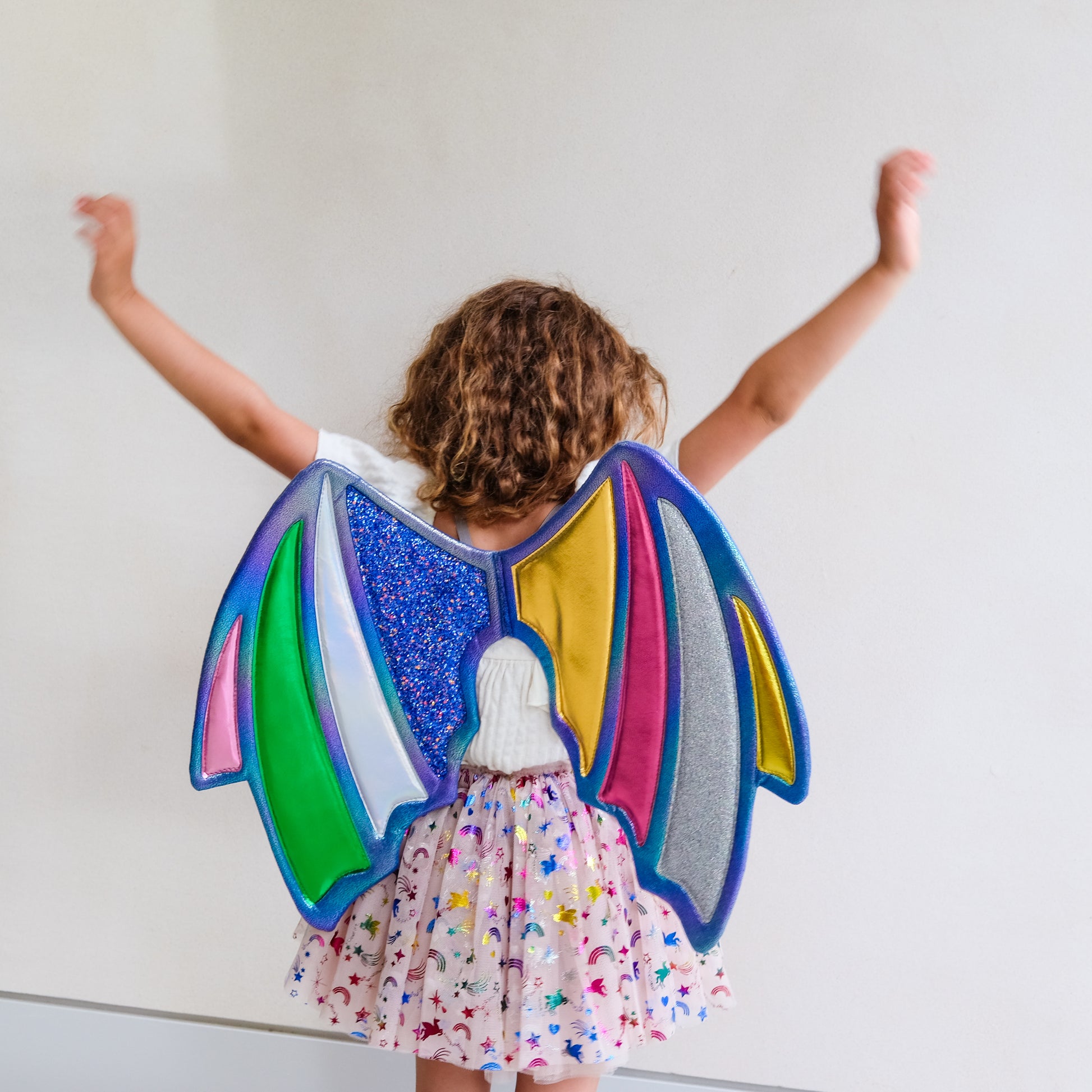 A child viewed from behind wearing a colorful patchwork angel wing costume over a white top and a pink star-patterned skirt, with arms raised against a plain light wall.