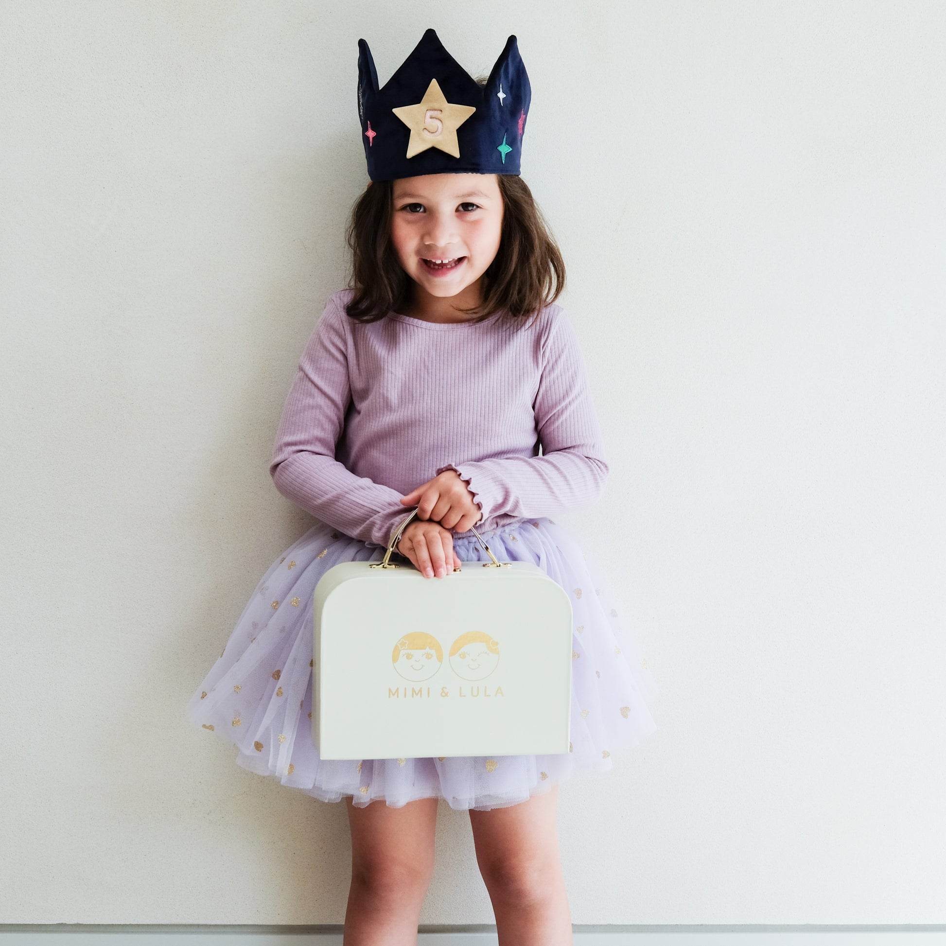 Child wearing a purple outfit with a blue velvet, star-patterned crown, holding a light green box.