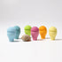 Six small egg-shaped containers in pastel blue, aqua, green, pink, orange, and yellow, arranged on a white studio background with a natural seashell placed in front of the center.
