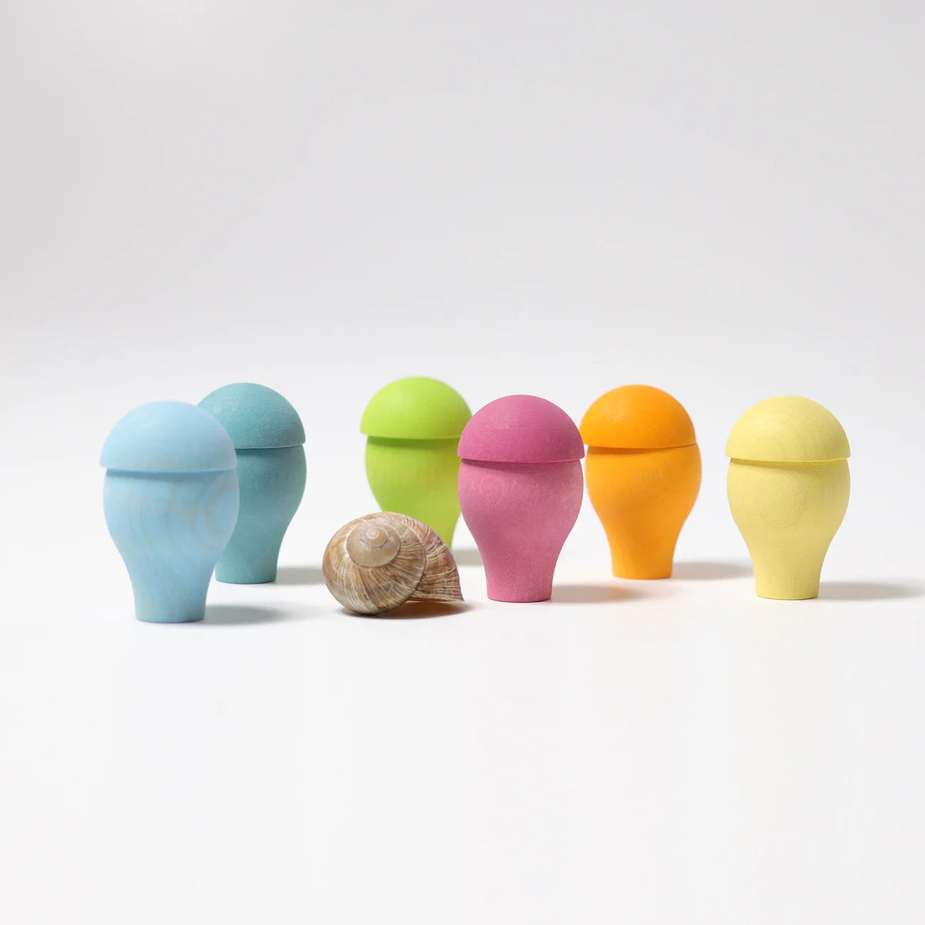 Six small egg-shaped containers in pastel blue, aqua, green, pink, orange, and yellow, arranged on a white studio background with a natural seashell placed in front of the center.