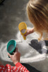 Two hands holding colorful plastic shovels and a bowl over sand on a beach.
