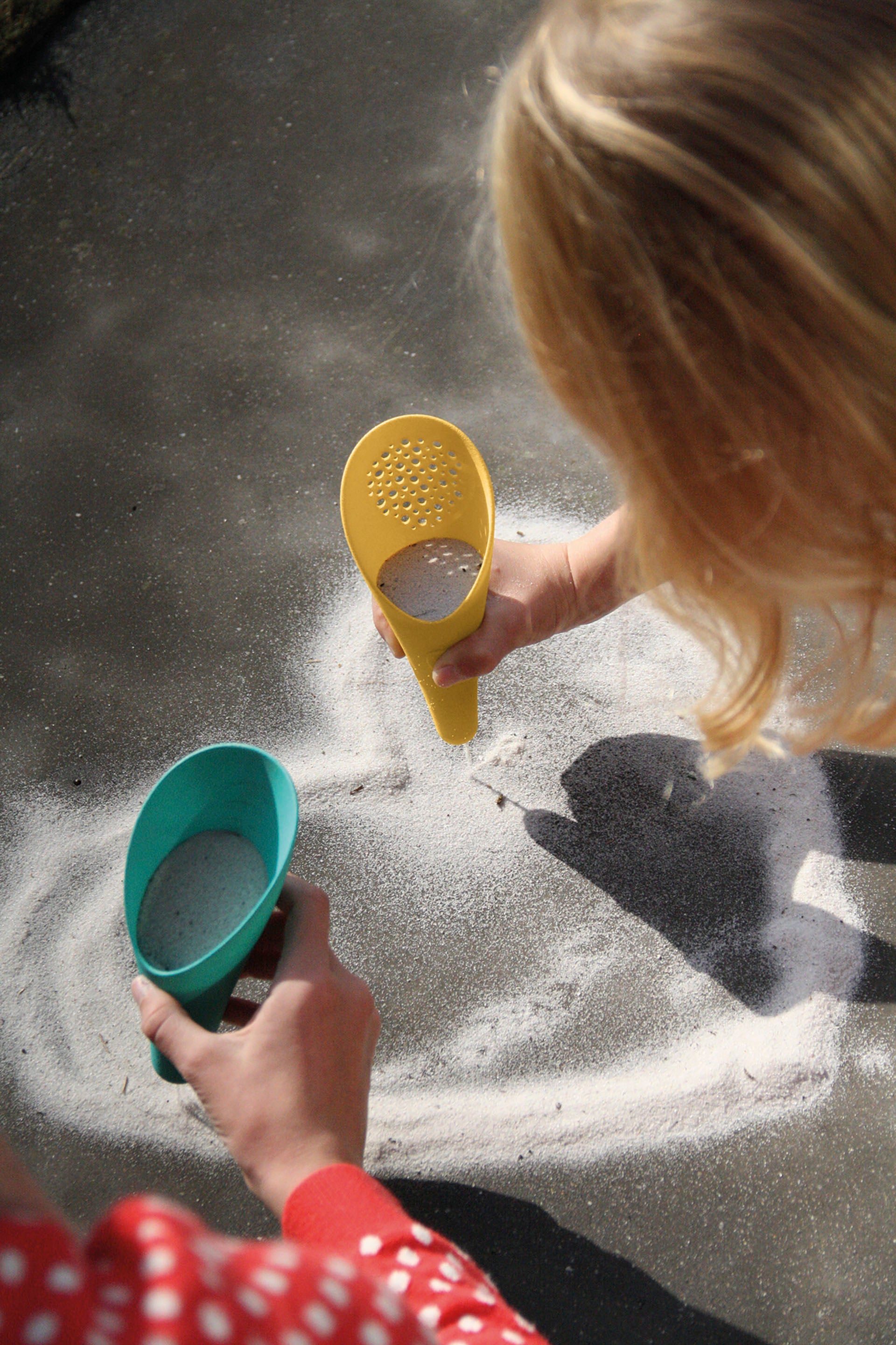 Two hands holding colorful plastic shovels and a bowl over sand on a beach.