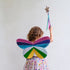 A child seen from behind wearing a colorful butterfly wing costume accessory, holding a glittery star wand against a plain light background.