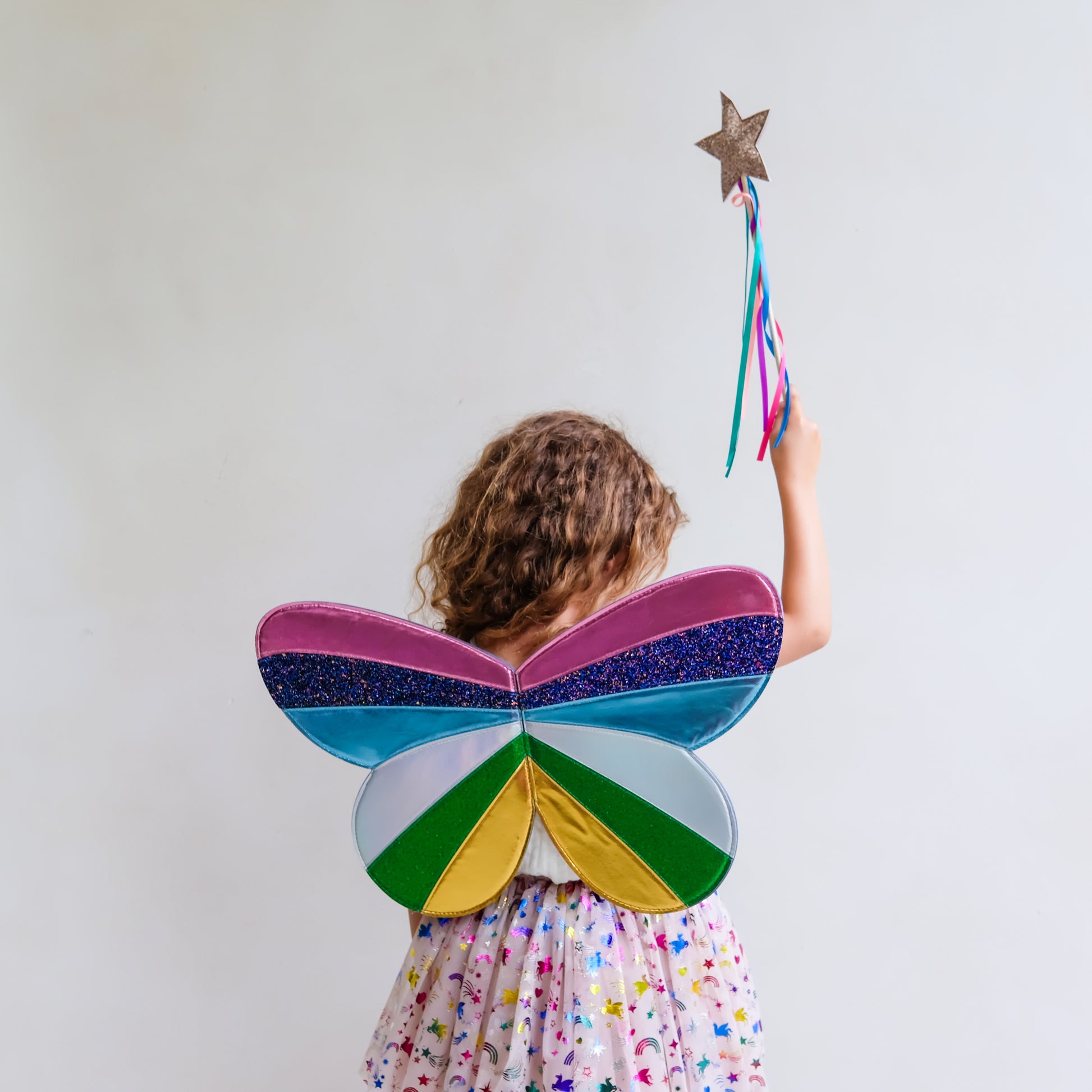 A child seen from behind wearing a colorful butterfly wing costume accessory, holding a glittery star wand against a plain light background.