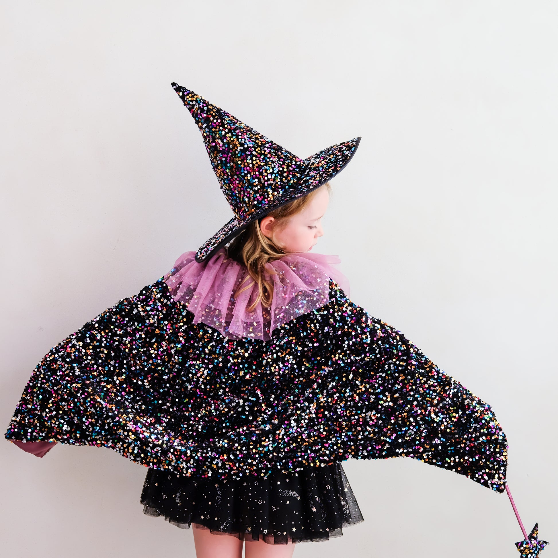 A young girl wearing a sparkly black witch costume with multicolored glitter, a pink tulle collar, and a matching pointed hat, photographed against a plain light background.
